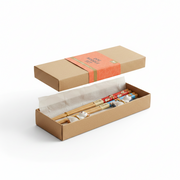 A person in a beige knit sweater holds open a brown cardboard gift box containing a set of chopsticks. The chopsticks have decorative animal-shaped handles and colorful bands, with a bright orange paper label on the box lid.