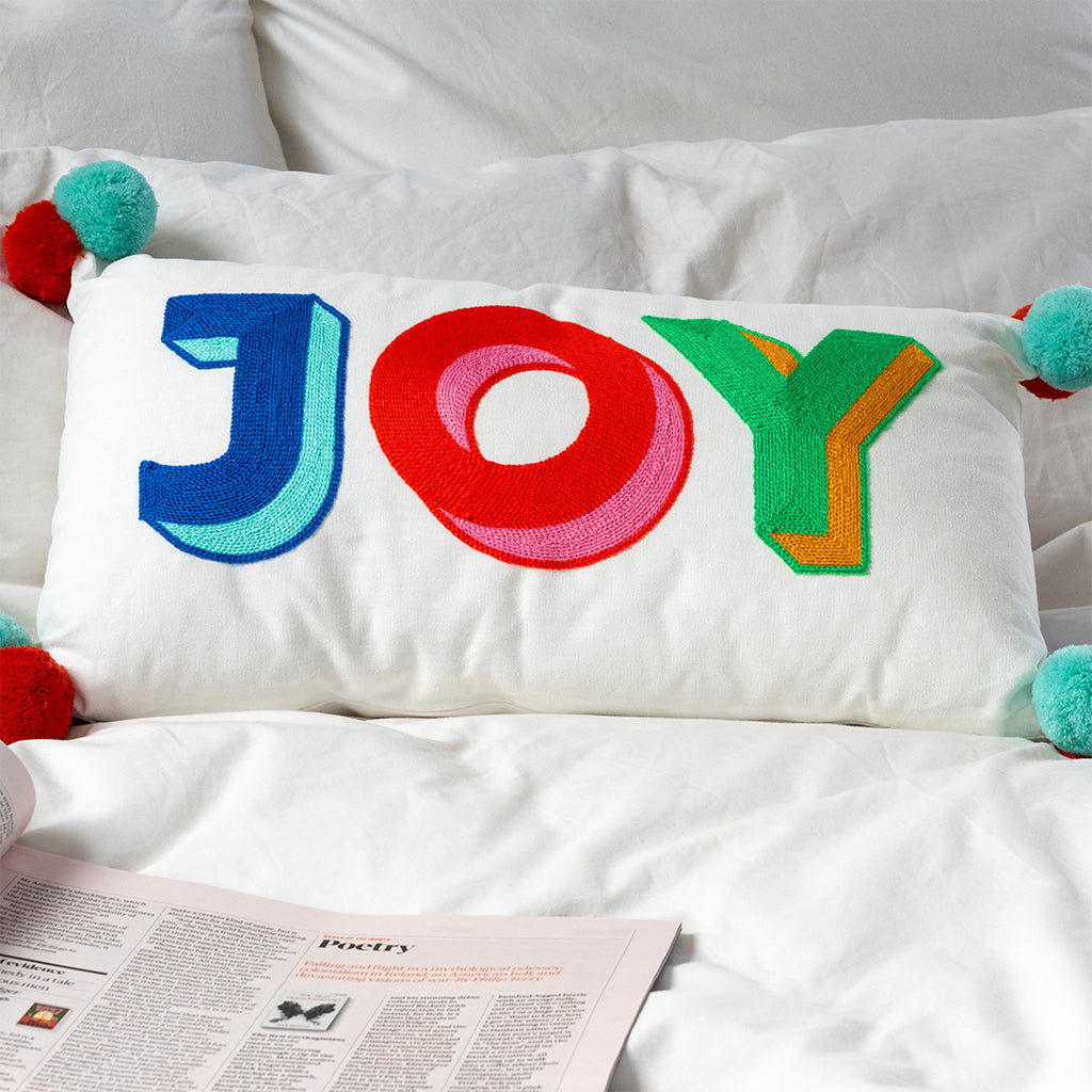 Joy pom pom cushion styled on white bed with pillows