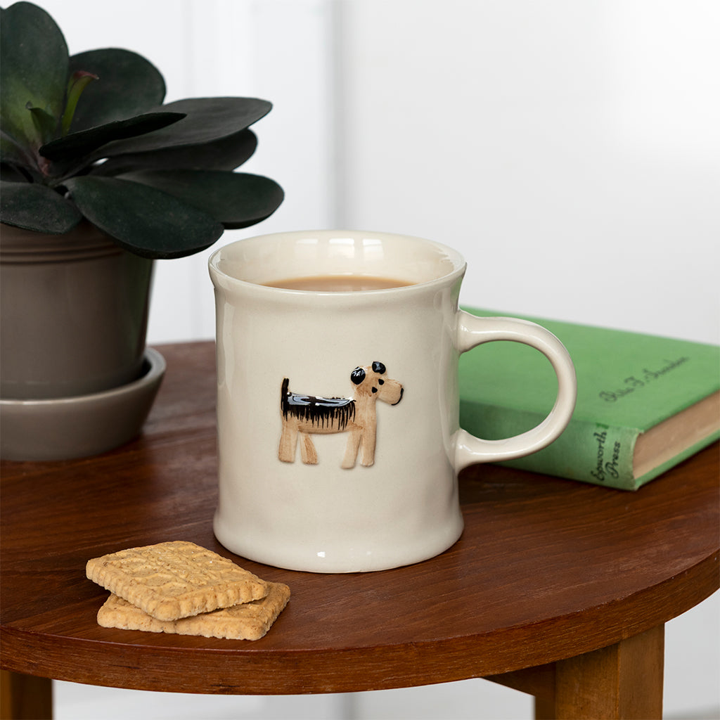 Cream dog mug on a wooden side table beside a biscuit, a book and a plant