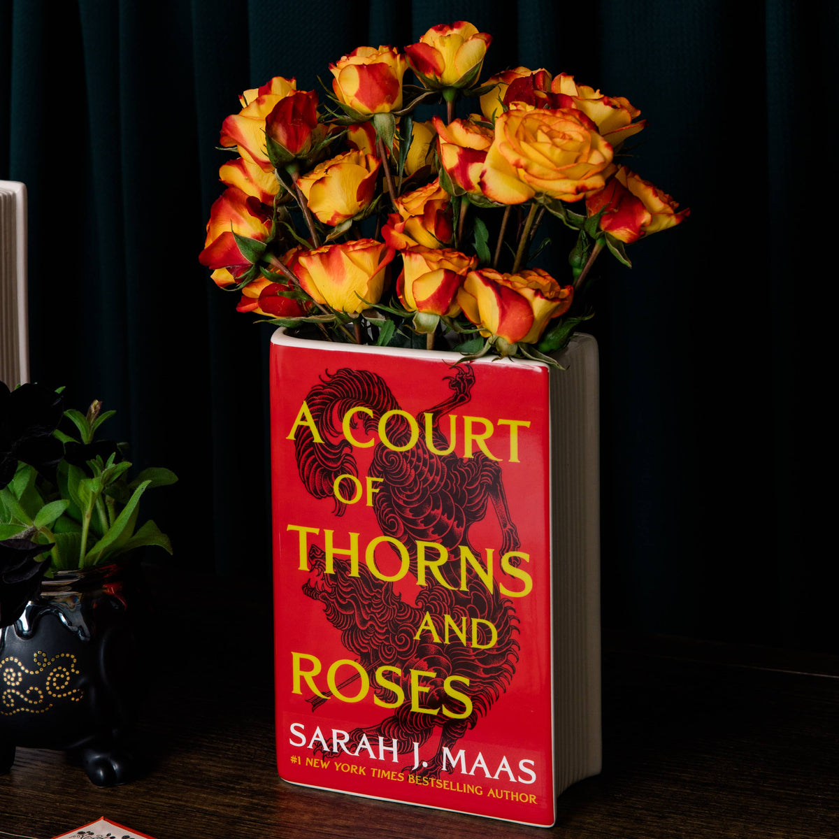 A Court of Thorns and Roses Book Vase holding orange roses on a dark table