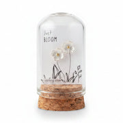 Small glass bottle with cork lid held by a hand, featuring a decorative label on a wooden surface.