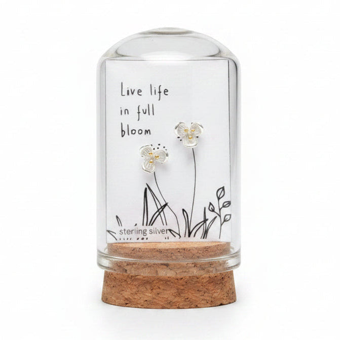 Small glass bottle with cork lid containing floral earrings and a label on a wooden surface.